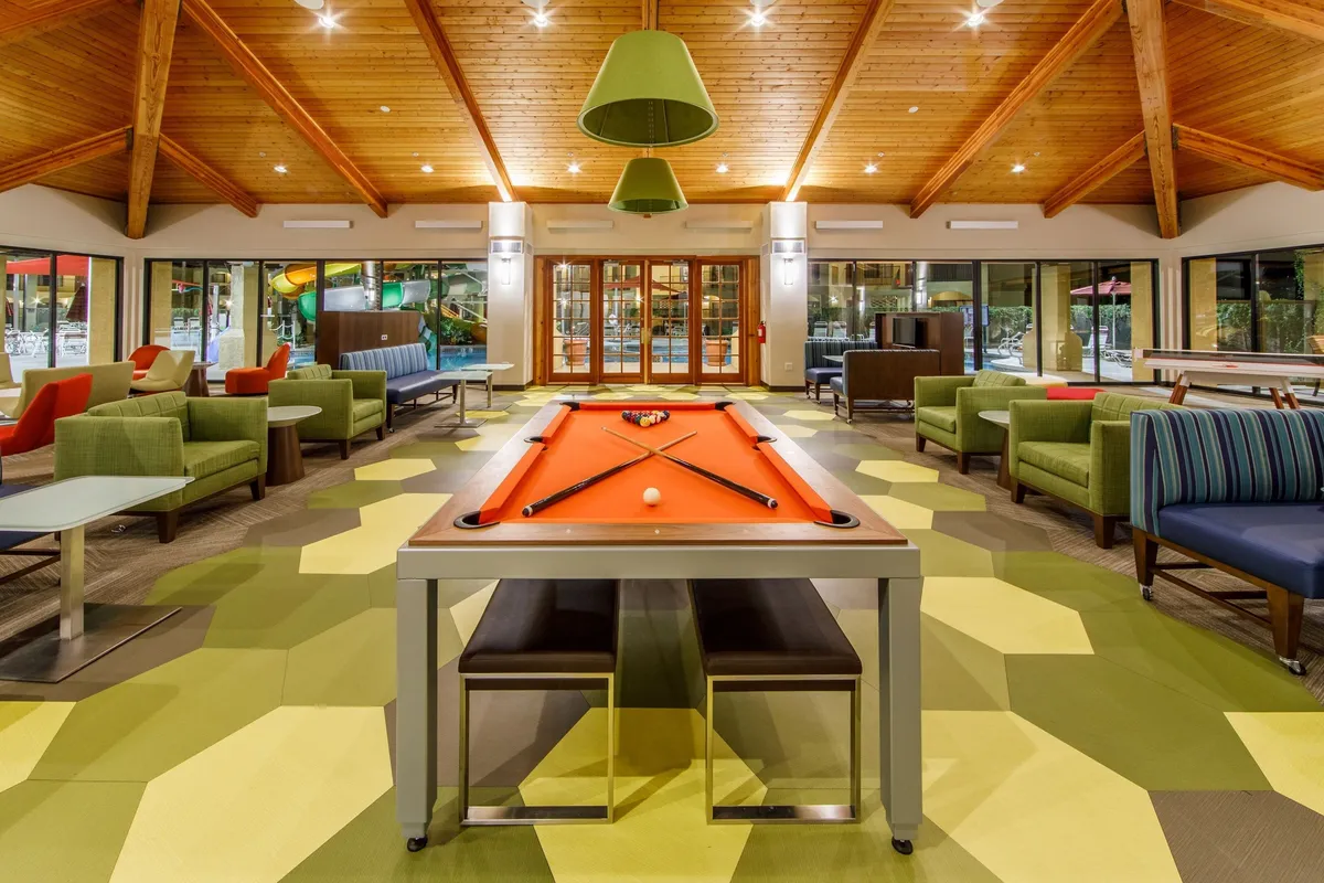 Inside, a lively game room is centered around a pool table, with green chairs for spectators and large windows providing natural light. The space suggests friendly competition, laughter, and leisure time for guests of all ages.