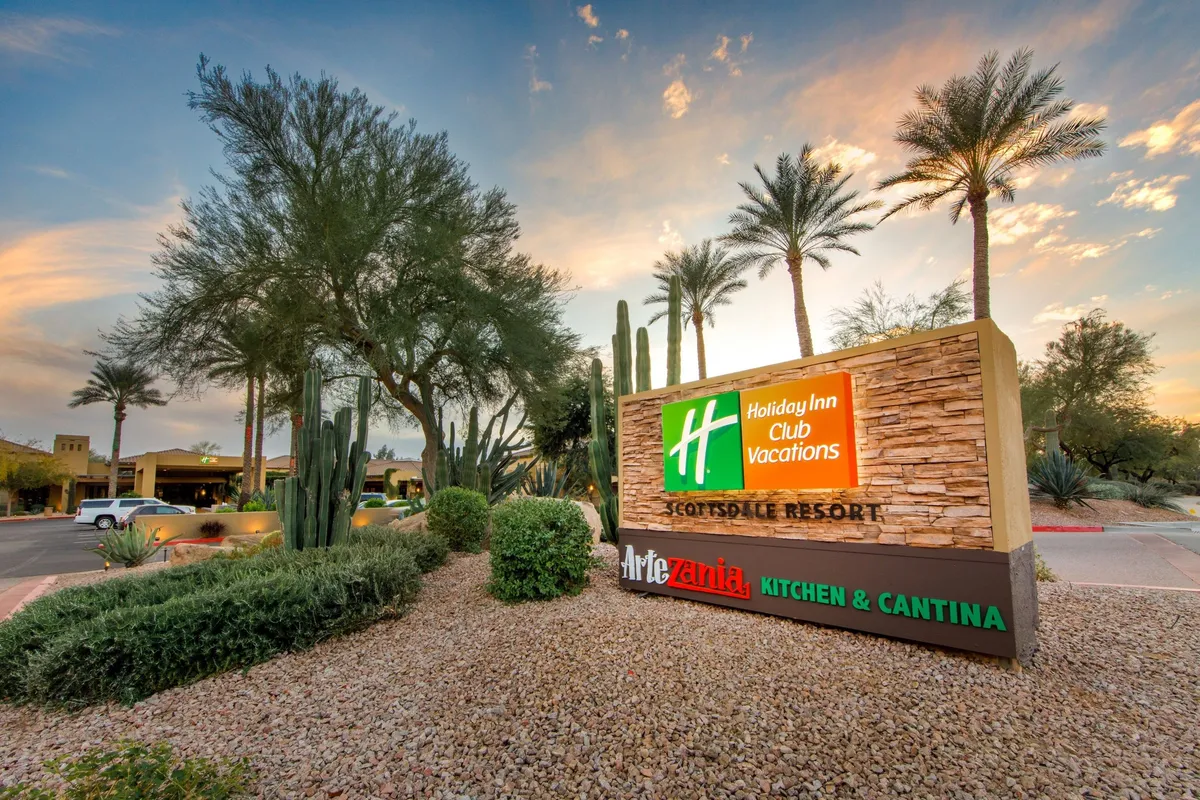 The image showcases the resort entrance framed by palm trees and cacti, with a clear Holiday Inn Club Vacations sign against a landscaped backdrop. The mood is serene and welcoming, hinting at the resort’s inviting atmosphere and proximity to outdoor adventures.