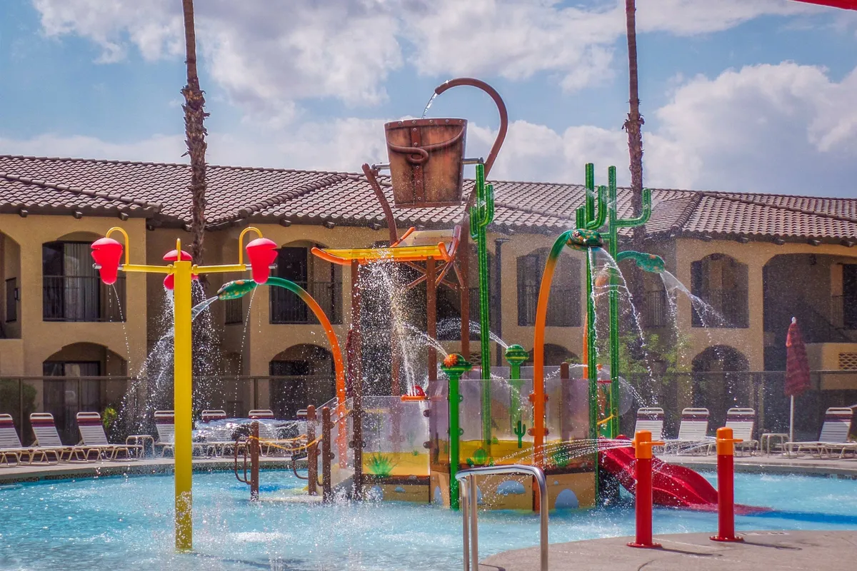 This image captures a vibrant outdoor pool area featuring a colorful water play structure with splashing water. Children are visible enjoying the water features, while families and groups relax around the pool, all under sunny skies. The atmosphere is lively and fun, perfectly illustrating family-friendly aquatic adventures.