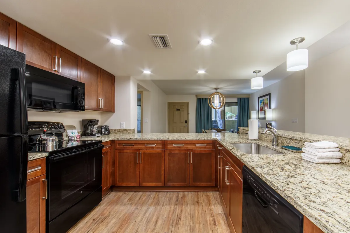 A warmly lit kitchen featuring granite countertops, dark wood cabinets, black modern appliances, and a spacious kitchen island. The setting invites group cooking, with enough space for multiple people to gather and prepare meals together.