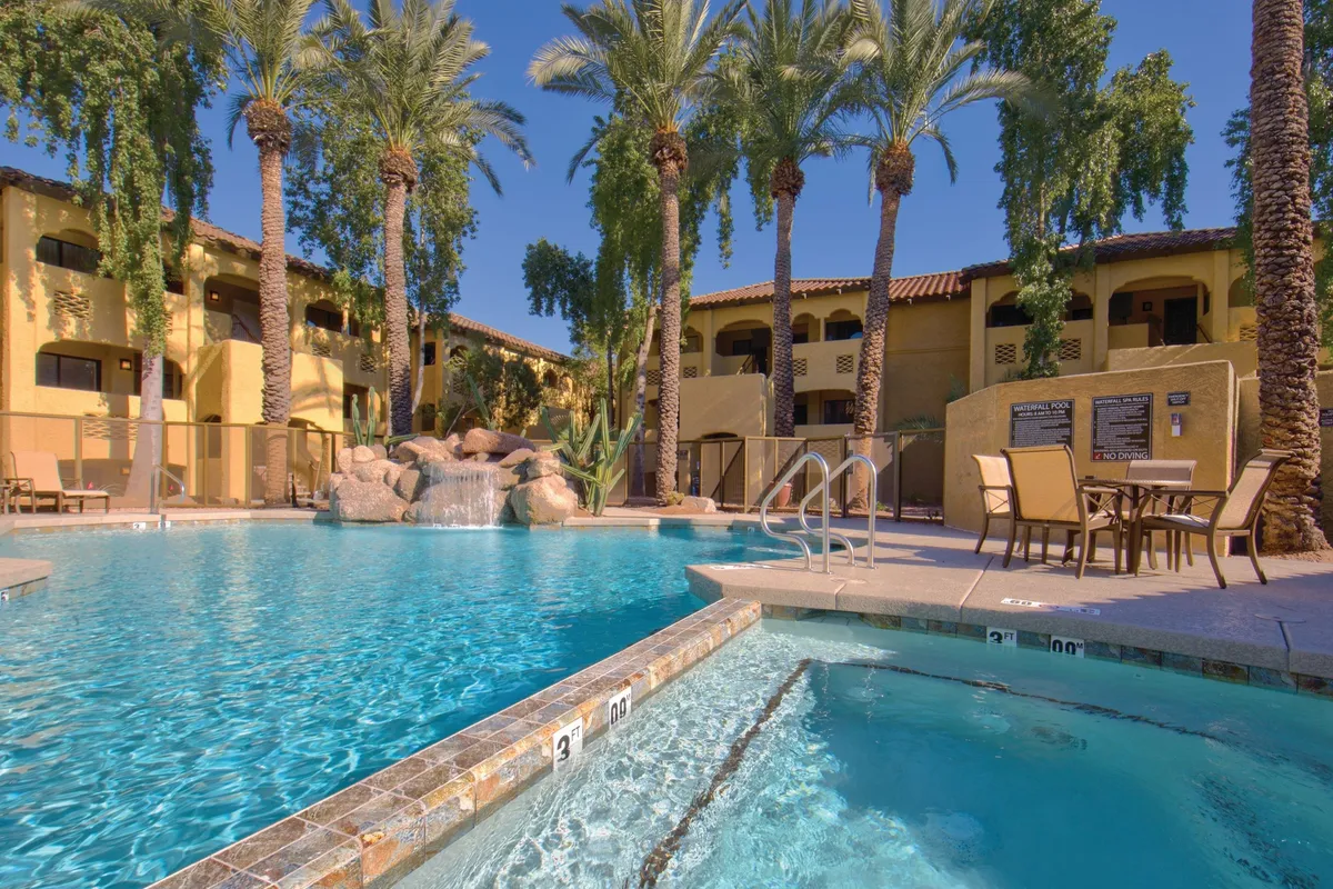 This image showcases a sparkling swimming pool surrounded by palm trees, loungers, and the facade of the resort building in the background. The pool features a serene atmosphere ideal for both family fun and tranquil relaxation, with guests enjoying sunbathing, swimming, and socializing.