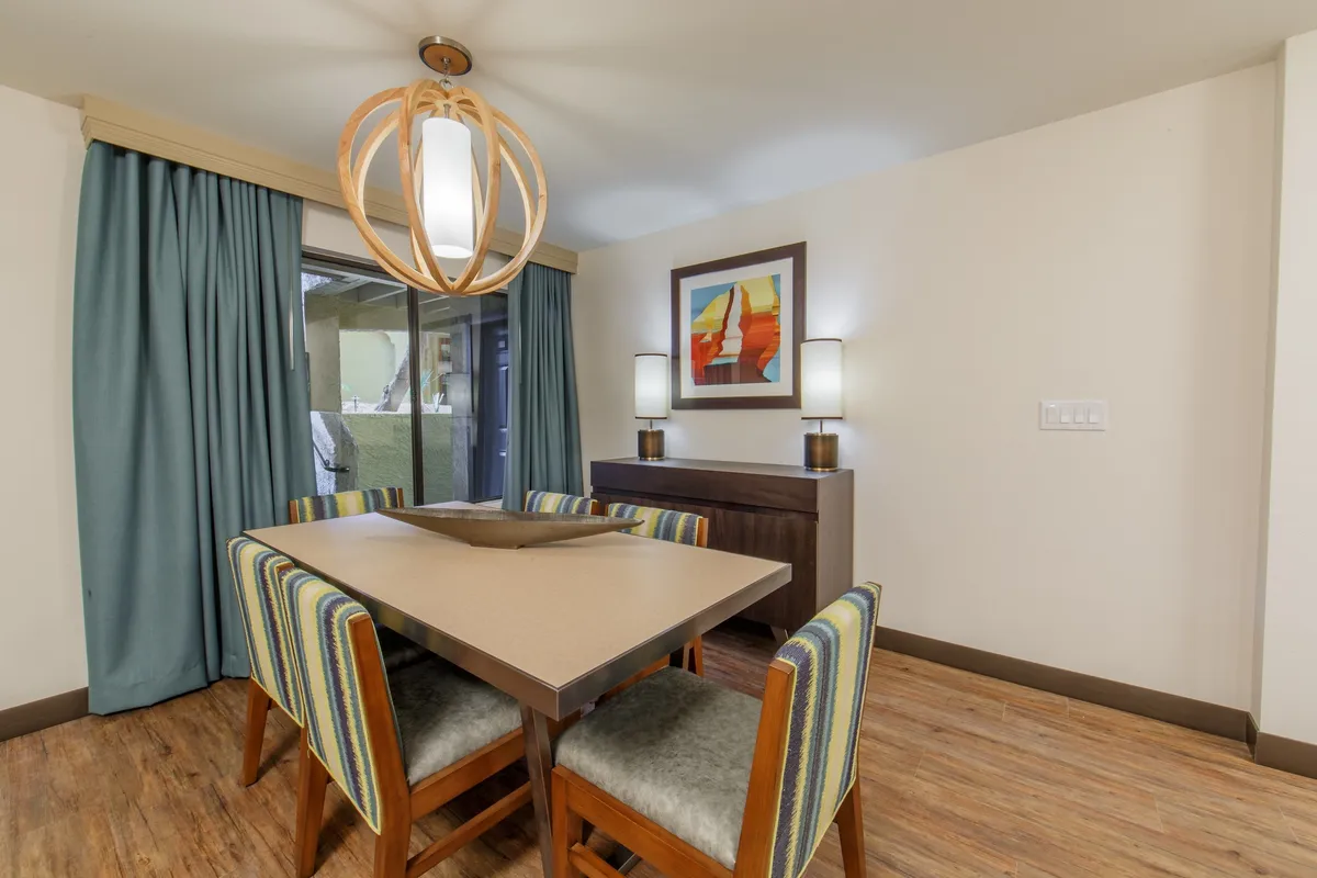 A cozy dining area centered around a wooden dining table beneath a stylish light fixture, surrounded by colorful artwork and soft curtains. The space feels intimate and welcoming, ideal for family meals or relaxed gatherings.