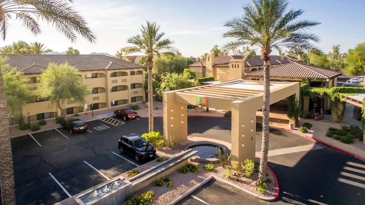The exterior entrance of the resort, featuring tall palm trees, an awning over the main doorway, a tranquil water feature, and a visible parking lot. The scene is both welcoming and practical, hinting at easy arrivals and access to amenities.