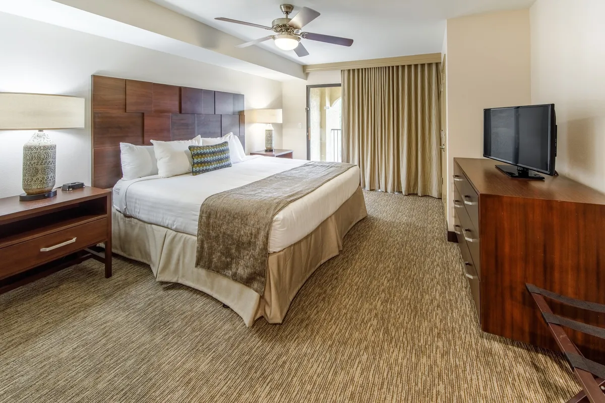 A serene villa bedroom featuring a king-size bed with wooden furniture, basking in natural light streaming through a balcony door. The space exudes comfort, offering guests a peaceful retreat to relax, watch television, or enjoy a quiet morning coffee while overlooking the scenery.