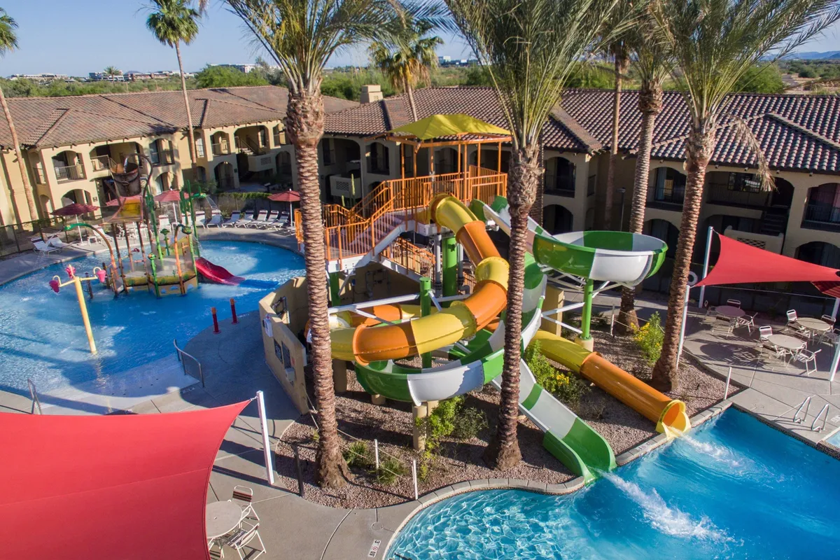 This vibrant exterior image features the resort's thrilling water slides, a swimming pool lined with palm trees and ample lounge seating. The setting pulses with family-friendly energy, showing kids and adults enjoying water play and soaking up the sun.